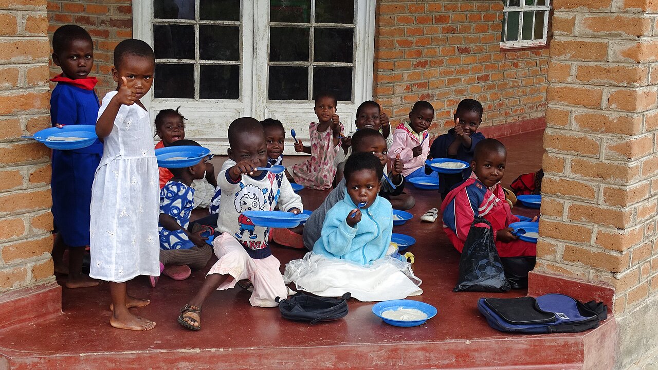Malawi: Auf einem überdachten Gang vor der Schule essen einige Kinder auf dem Boden sitzend.